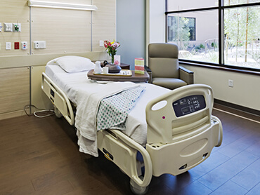 View of patient room with bed and chair next to a large window.