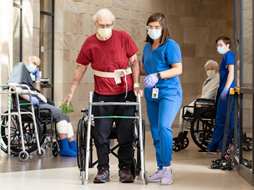 Female therapist in blue scrubs walks with older male patient in red shirt using a walker.