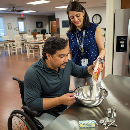 Patient in a wheelchair is pouring food into a bowl with the support of his occupational therapist